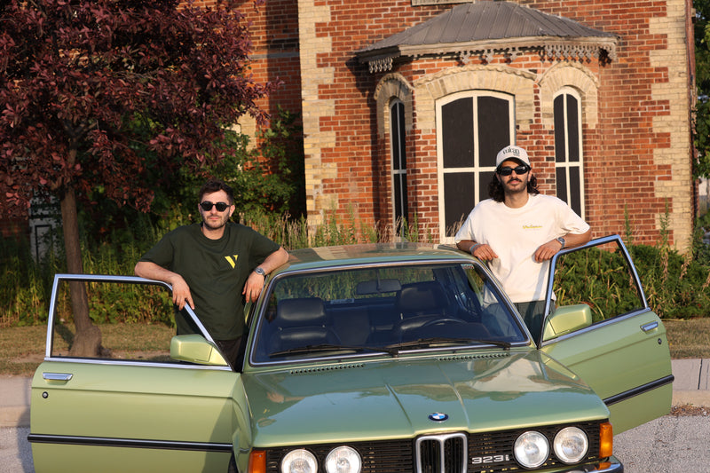 Two men standing next to a green BMW car in front of a brick building.